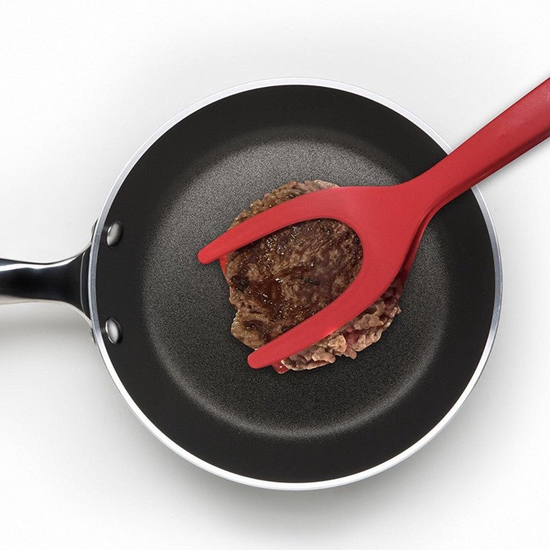 Red 2-in-1 spatula tongs flipping a burger in a non-stick pan. Kitchen cooking utensil.