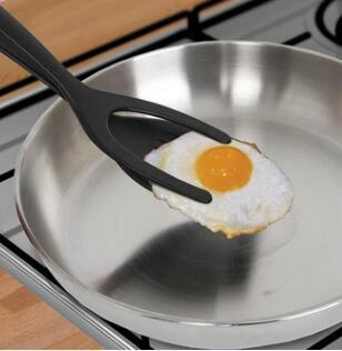 Black 2-in-1 spatula tongs flipping a fried egg in a skillet. Kitchen utensil.