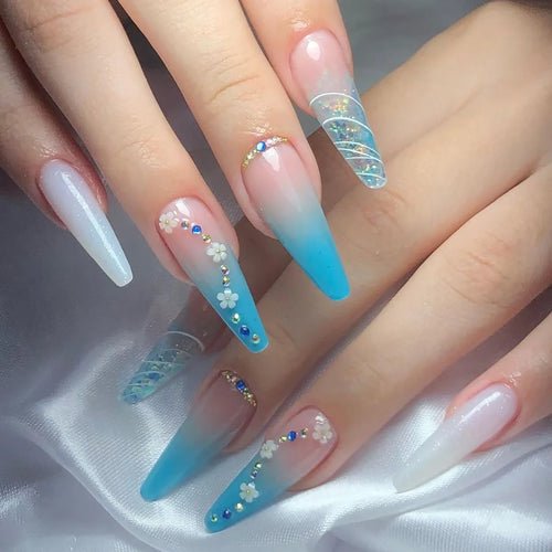 Long coffin-shaped false nails with a blue ombre, floral, rhinestone, and glitter swirl design.