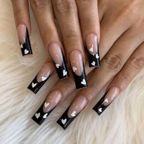 Long coffin-shaped black false nails with white heart details and French tip design.
