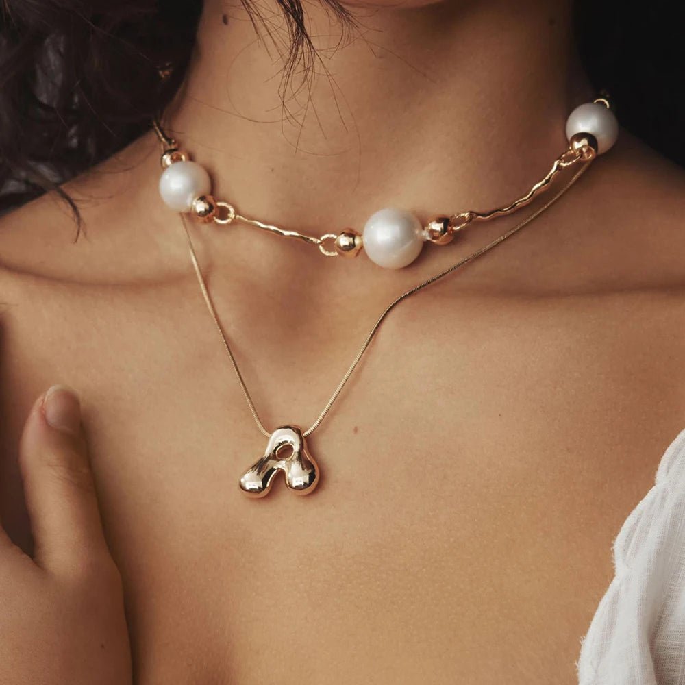 Model wearing layered gold necklaces: a pearl beaded choker and an initial pendant necklace.